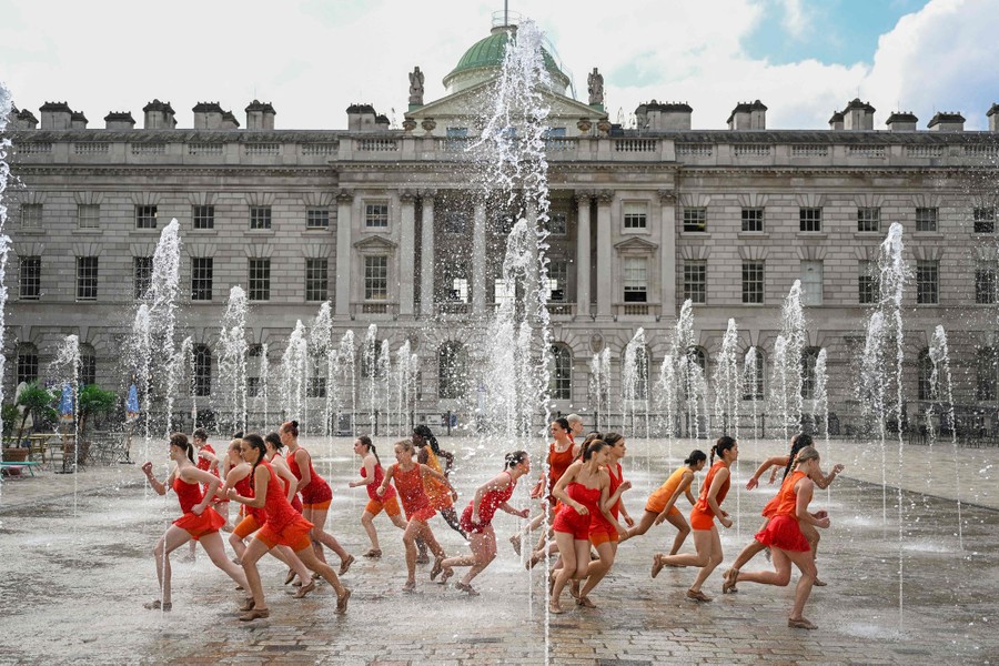 A couple dozen performers wearing red dresses run in multiple directions in a public fountain.