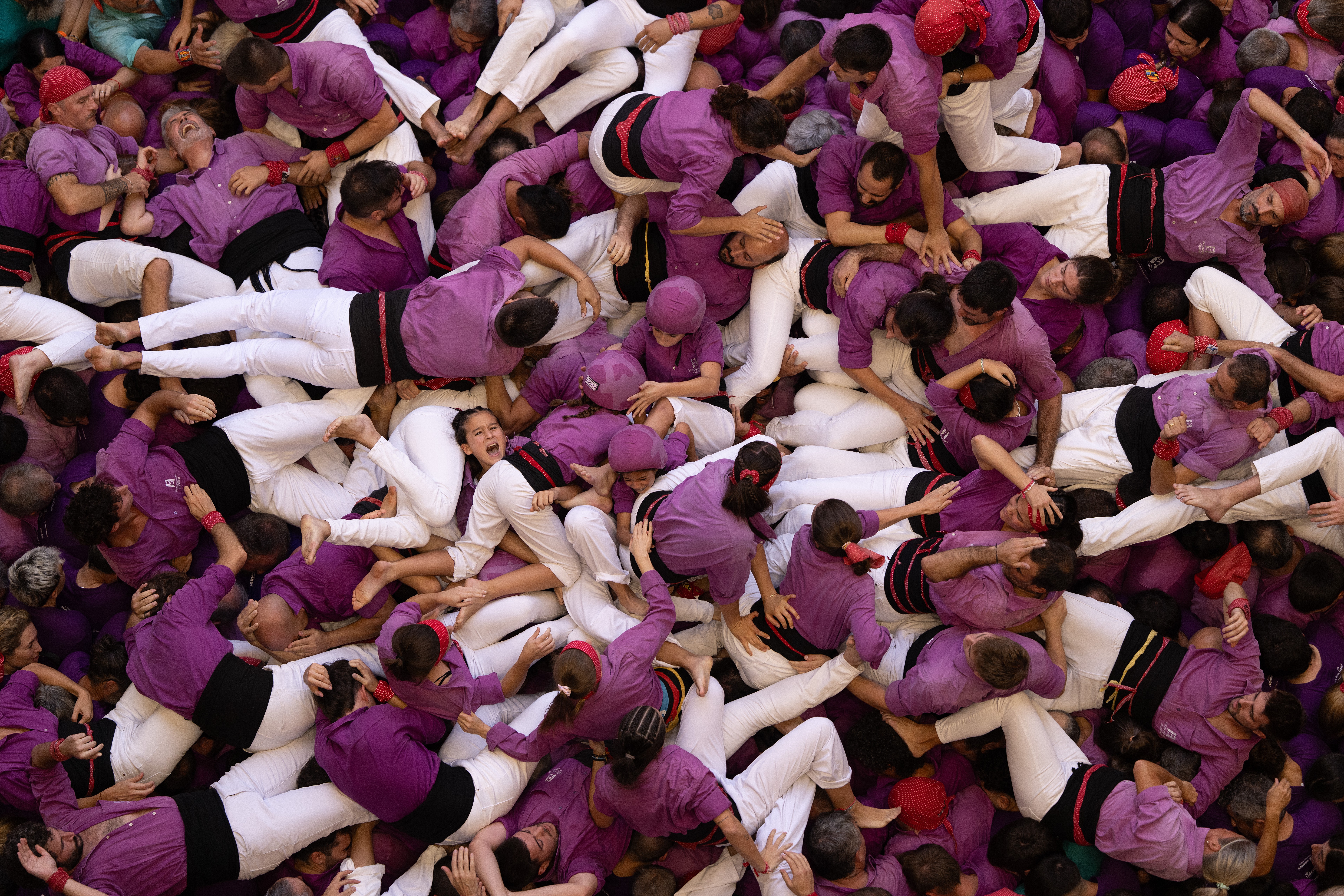 An overhead view of a team of people wearing purple and white uniforms, piled on top of each other after a fall while they were attempting to build a human tower