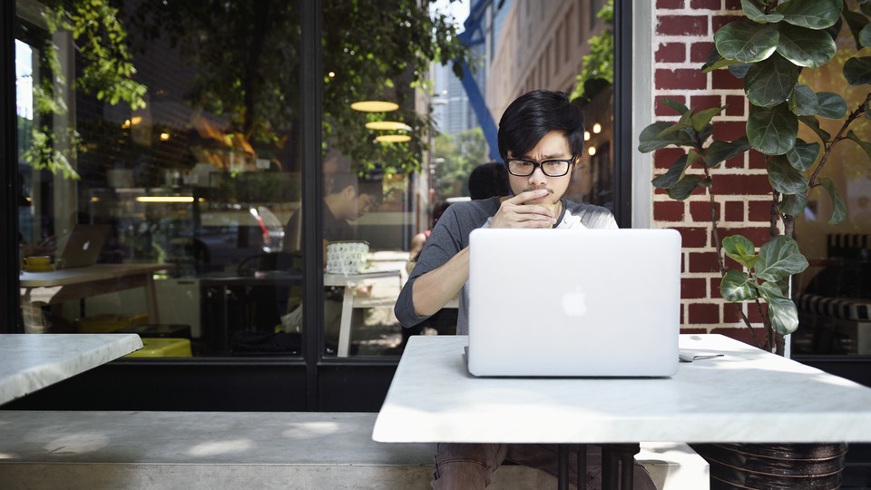 A young man looks thoughtfully at a laptop while sitting at a cafe