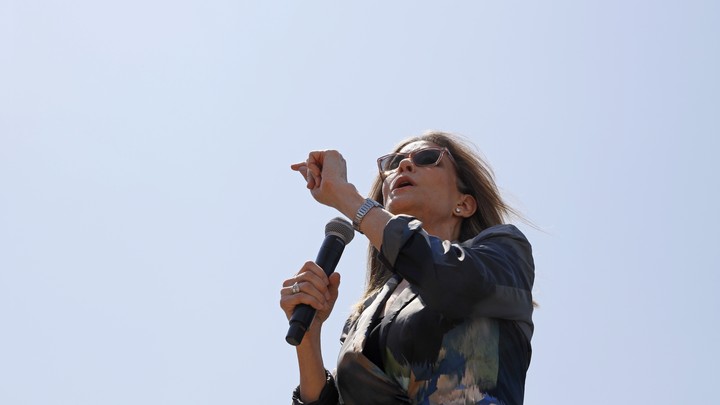 Marianne Williamson speaks into a microphone at the Iowa State Fair. She's wearing sunglasses and a watch, and the blue sky is visible behind her.