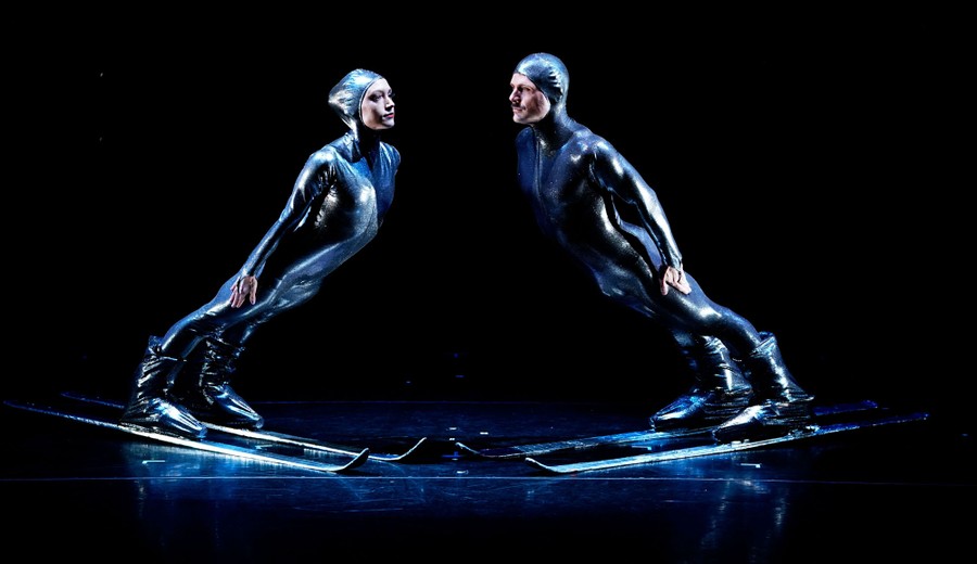 Two performers in full body suits lean forward while wearing skis on a stage.