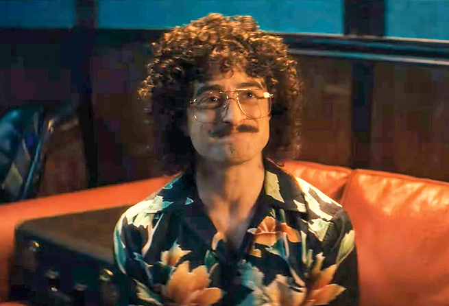 still image from movie with Radcliffe in curly wig, gold-framed glasses, and mustache, wearing Hawaiian shirt as Weird Al