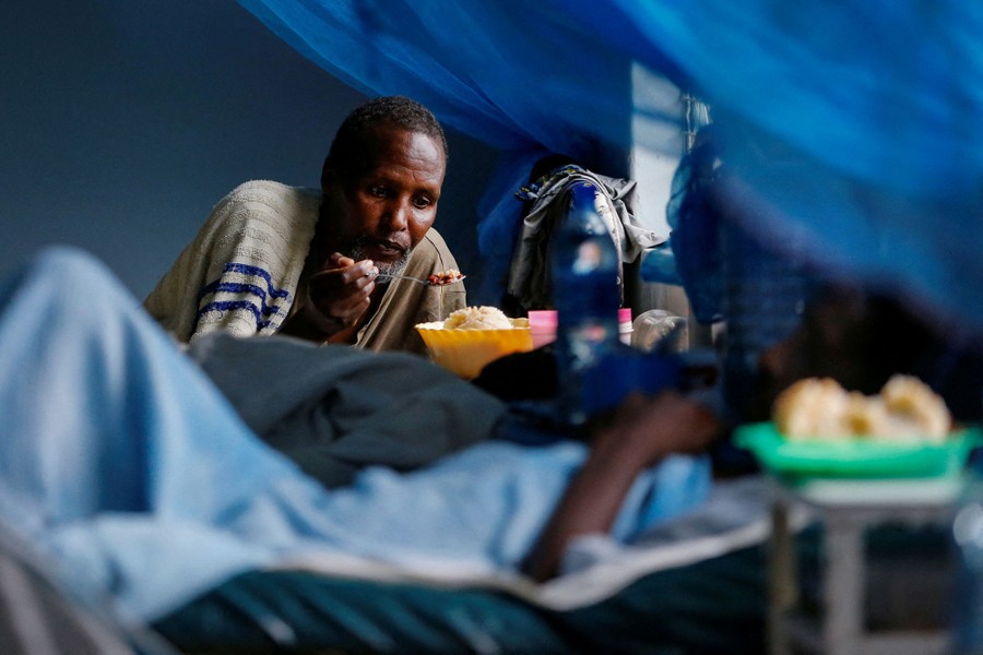 A person holds a spoon as they feed another person who is lying down in a hospital bed.