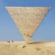 A pyramid balances on its point, upside down, in the desert with blue sky and 3 small figures