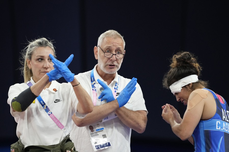 Two doctors wearing rubber gloves cross their arms in X shapes, as they disqualify an injured wrestler, who reacts nearby.
