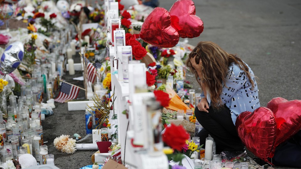 A woman crouches, head in hand, in front of a makeshift memorial near the scene of a mass shooting in El Paso, Texas. The memorial is made up of white crosses, prayer candles, American flags, and red heart-shaped balloons, among other items.