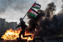 A man holding a Palestinian flag runs in front of a blazing fire.