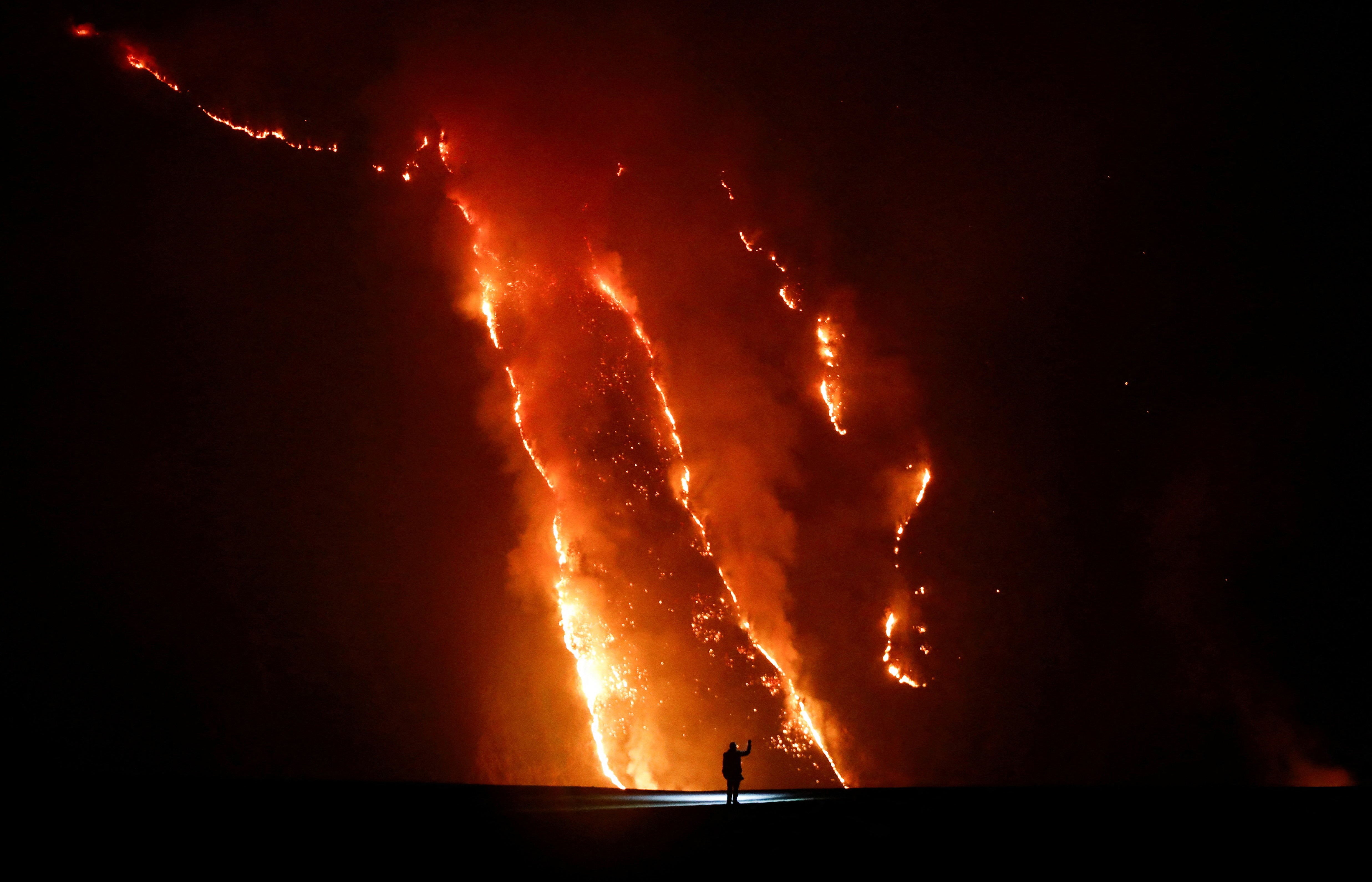 A person observes a wildfire burning across a hill slope at night.