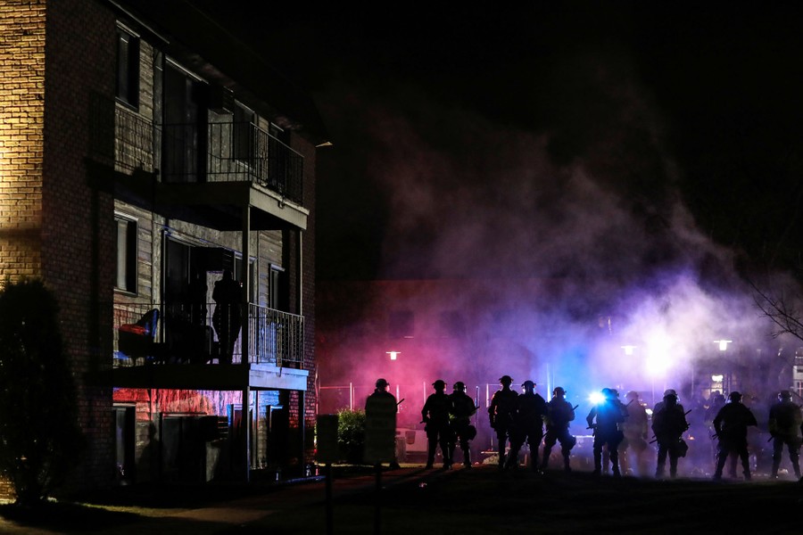 Riot police move forward at night, with smoke rising above.