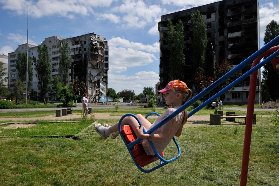 A girl rides a swing on a playground in front of several destroyed residential buildings.