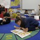 A girl crouches over a book on a colorful rug. Other children around her also have books on their laps.