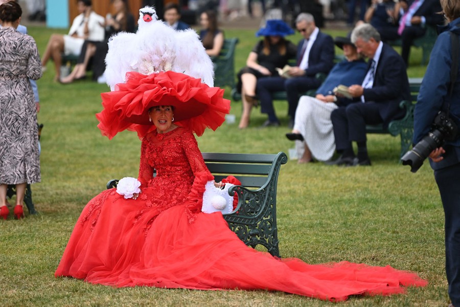 A woman sits on a bench, wearing a long red gown and a huge hat with a stuffed swan on top of it.