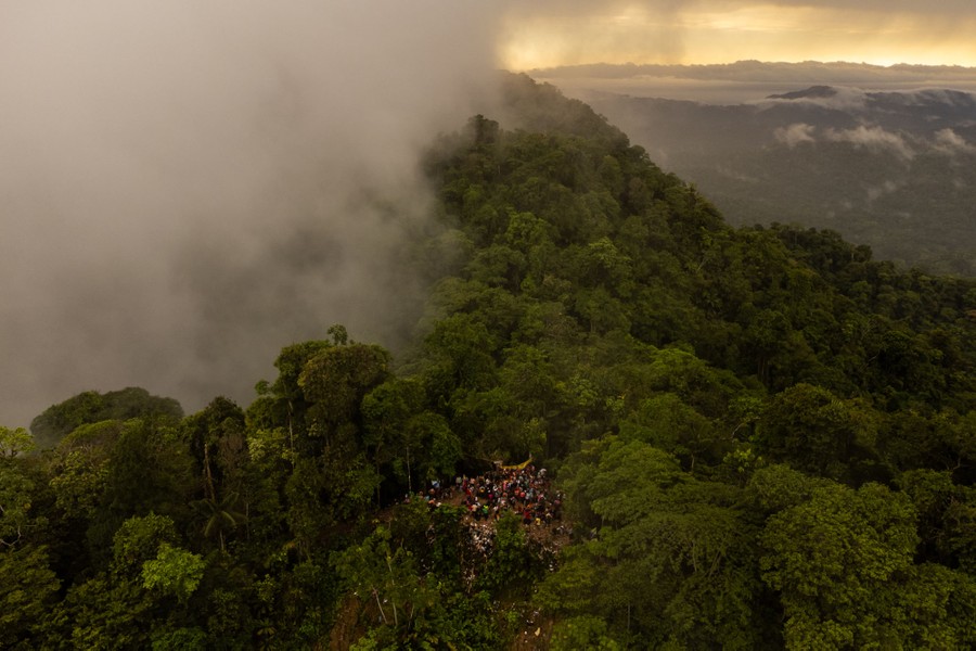 An aerial view of a group of people standing in a small clearing in a dense jungle on mountainous terrain