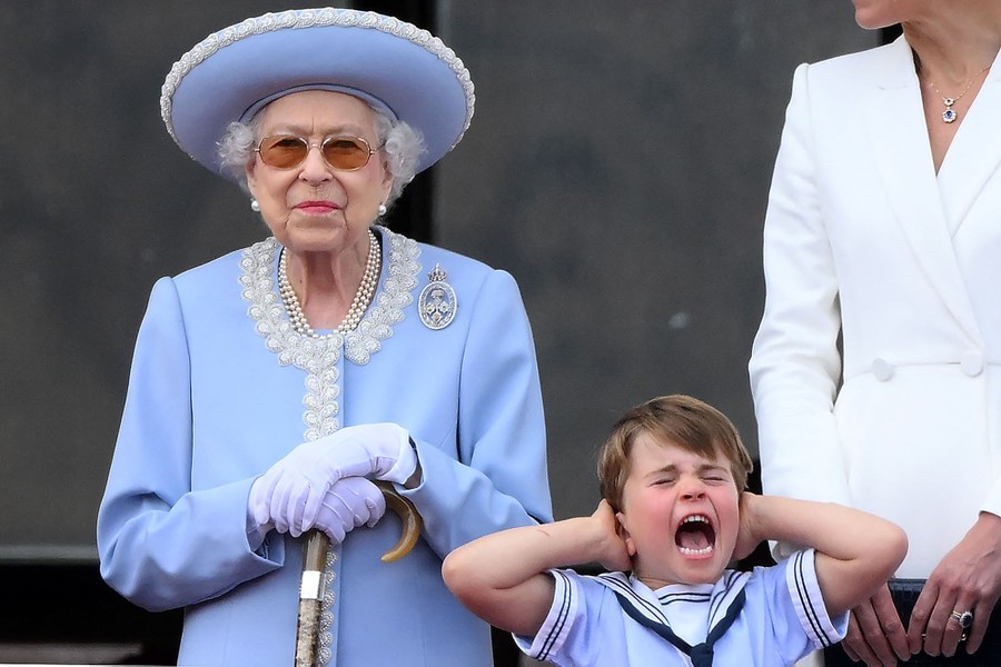 A young boy stands beside Queen Elizabeth II on a balcony, shouting, with his hands over his ears.