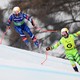 Two downhill skiers race down a course, the one in front, acting as a guide, wears a bib with a large letter "G."