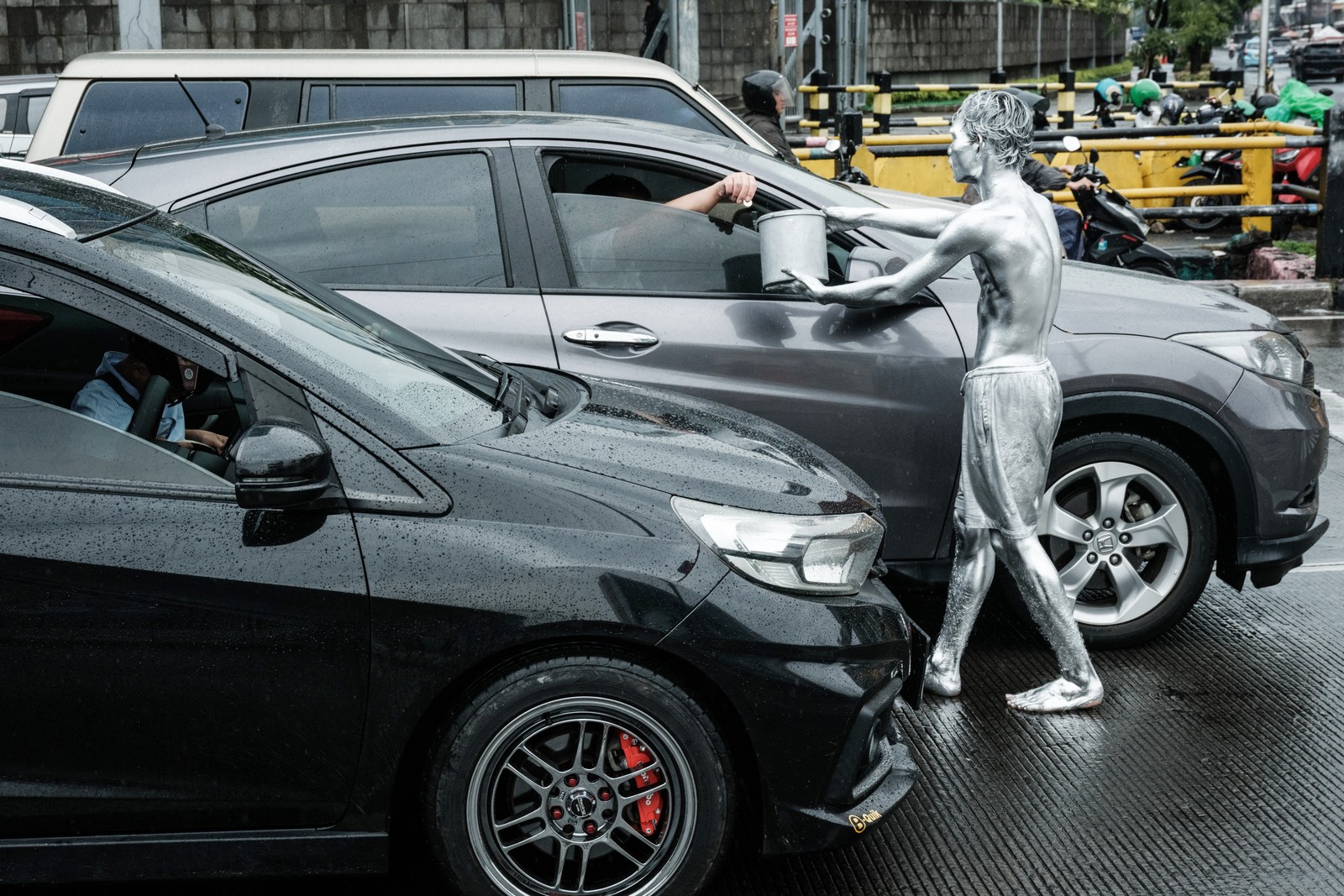A street performer covered in silver paint holds out a bucket for tips beside a car.