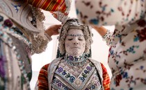 A bride, her face covered in white makeup lined with patterns, has two people help with her traditional clothing.