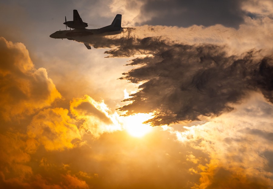 An aircraft drops a plume of water against a backdrop of the sun behind clouds.