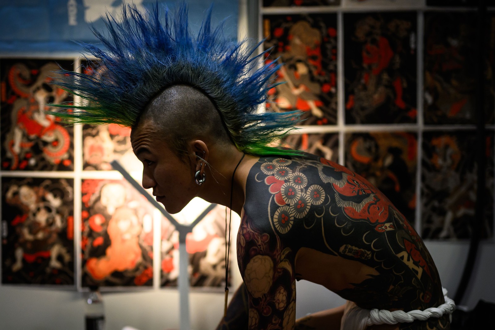 A heavily-tattooed man with a blue mohawk sits beside a wall covered in colorful prints.