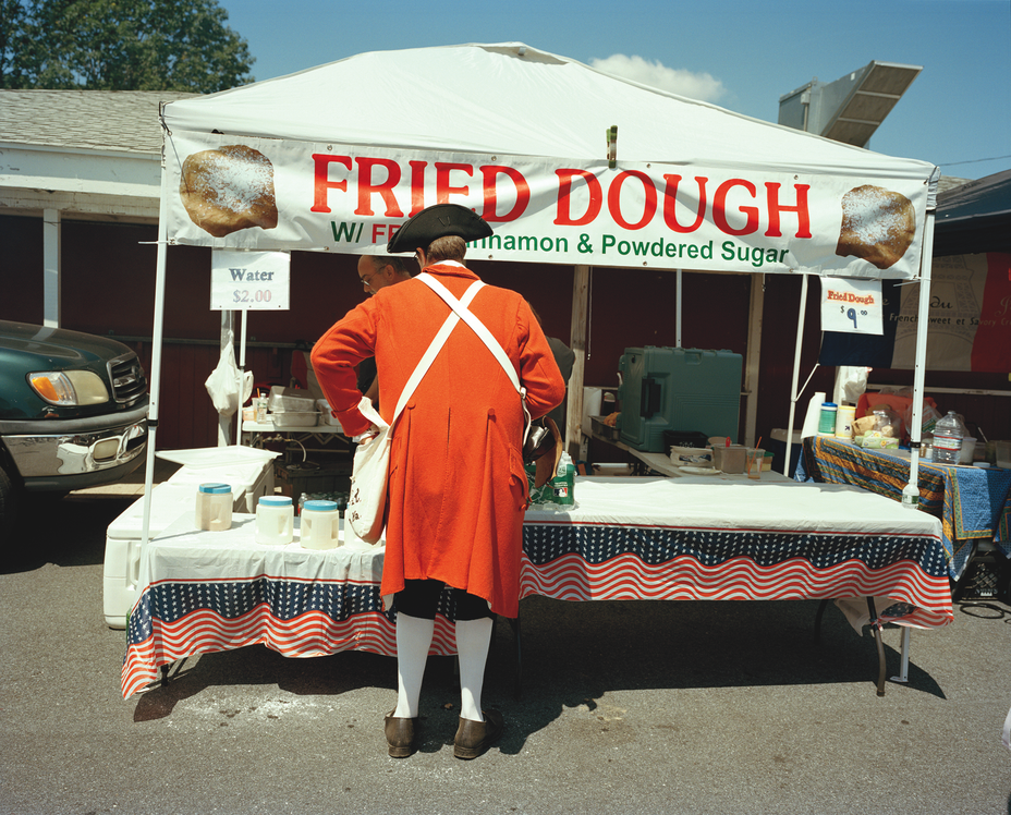 photo from behind of a reenactor playing a British soldier dressed in full regalia ordering food at a Fried Dough tent in the parking lot of the reenactment