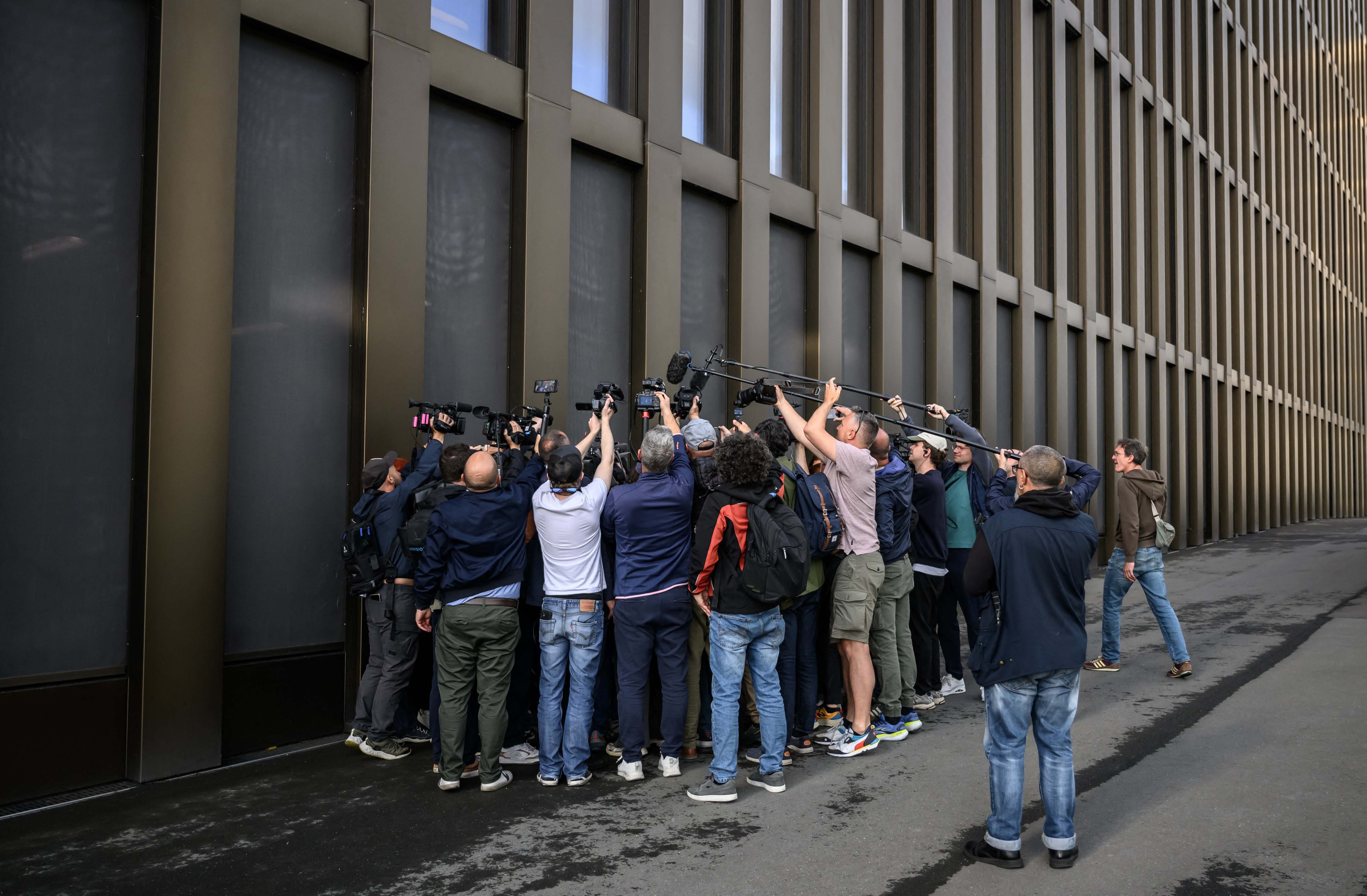 A tight group of journalists surround a lawyer, crowded against the outside wall of a building.