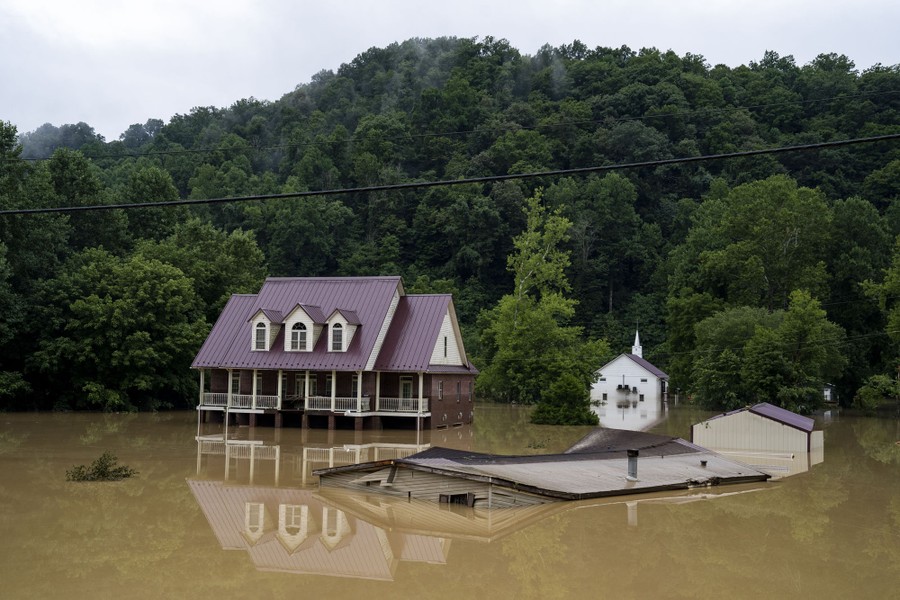 Several structures are seen beside a tree-covered hill surrounded by muddy floodwater.