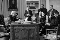 A black-and-white photograph of President Donald Trump on the phone while meeting with senior officials in the Oval Office.