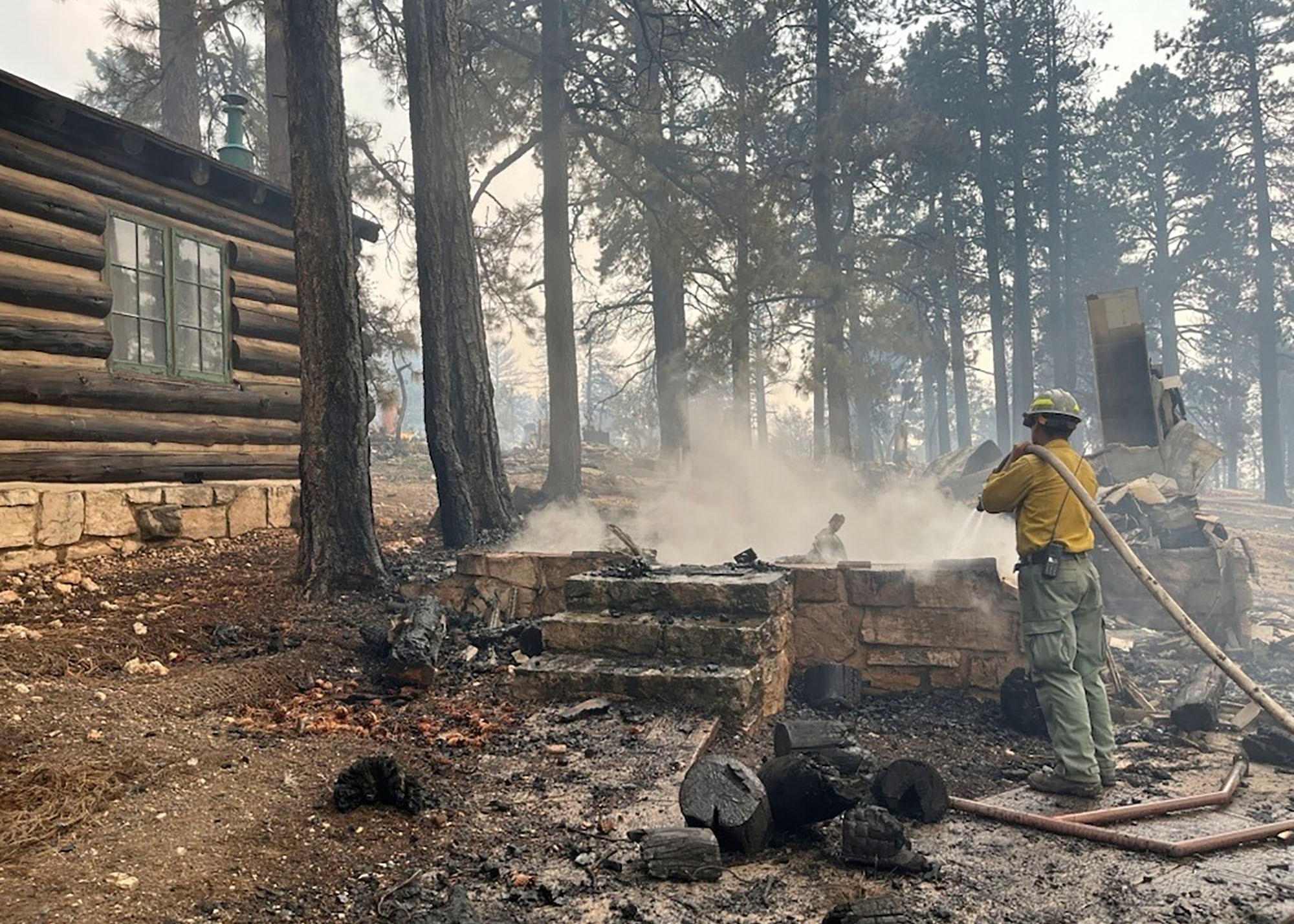 A firefighter works at the charred remains of a building in a national park, beside a log building.