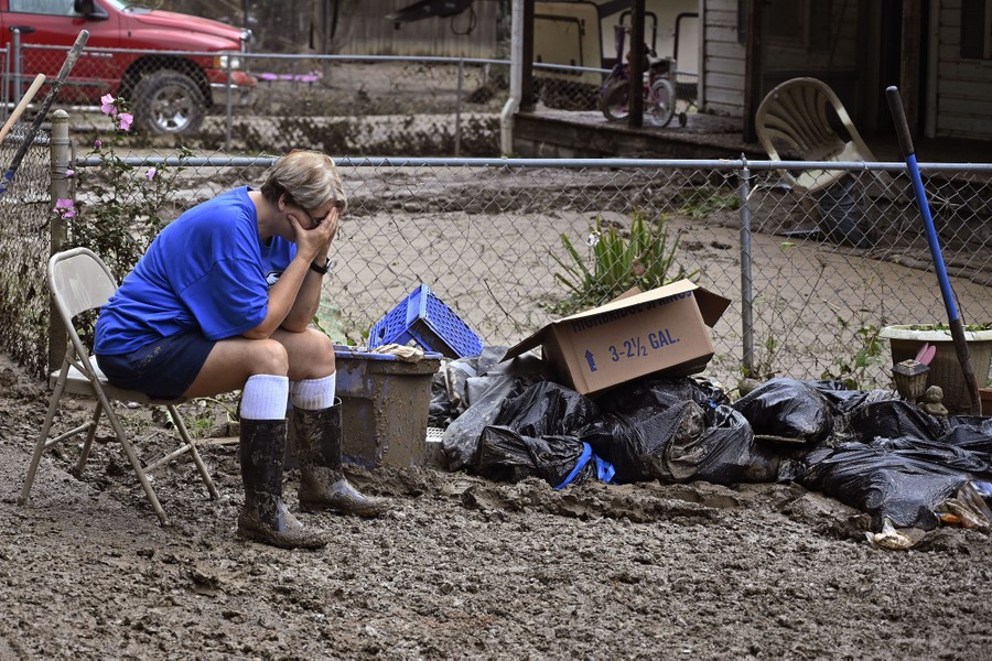 A person sits on a folding chair in a yard covered with mud, surrounded by boxes and garbage bags.