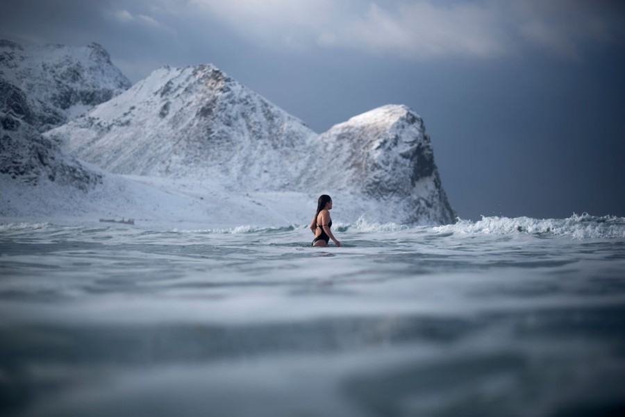 A person in a swimsuit wades into the surf, with snow-covered mountains in the background.