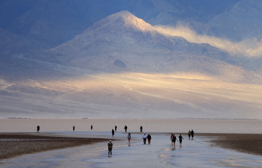 More than a dozen people walk in and along shallow water in a broad desert basin, with mountains in the background.