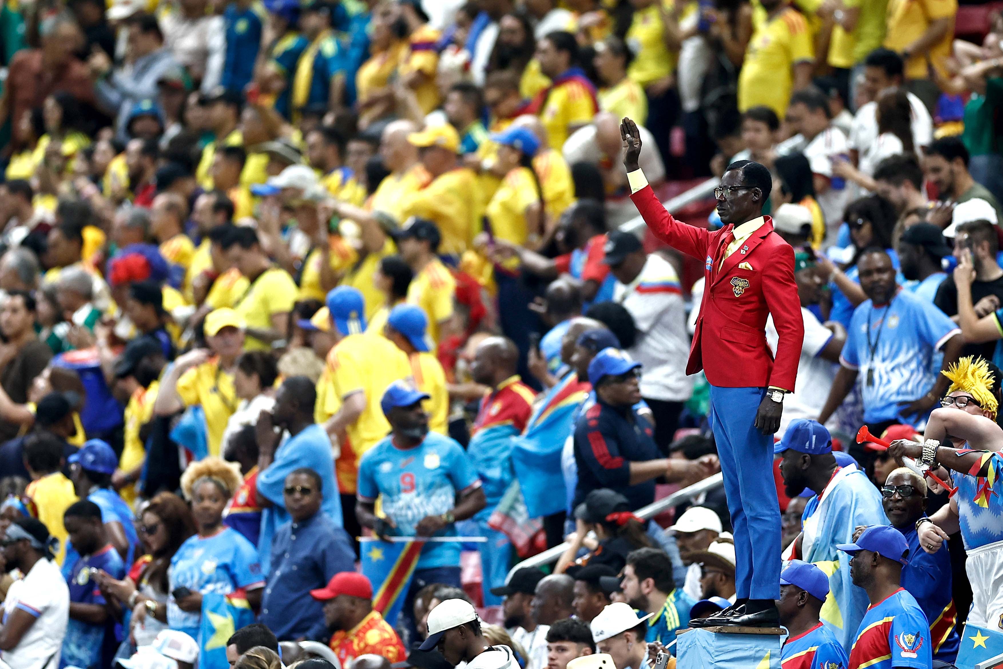 A football supporter stands stiil, bolt-upright, one arm raised, among a stadium crowd during a game.
