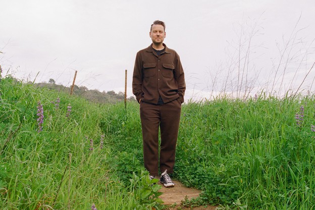 Color photo of man in brown shirt and pants standing with hands in pockets on trail surrounded by green grass and wildflowers.