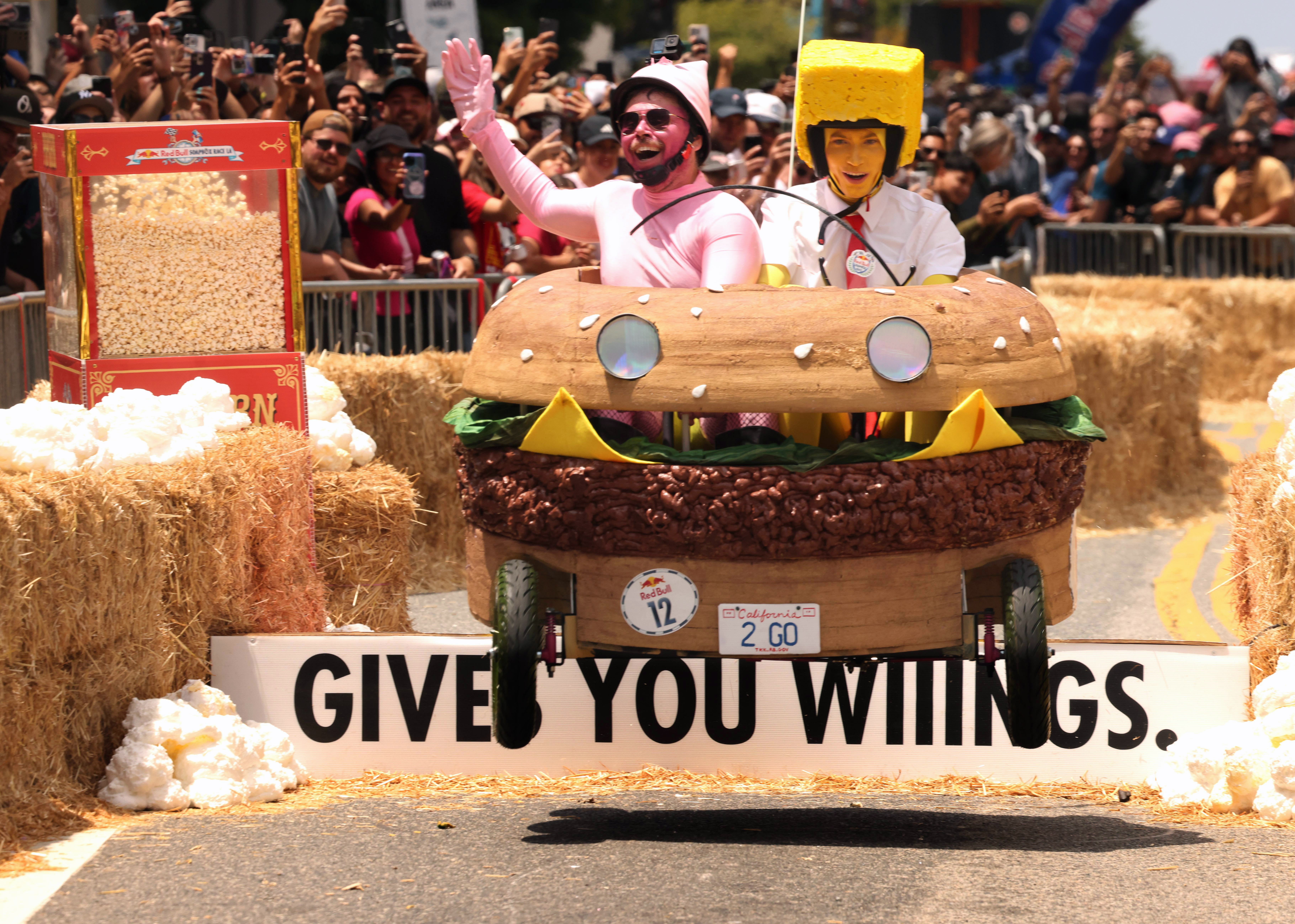 Two people in costumes race a hamburger-shaped soapbox-derby cart, making a jump.