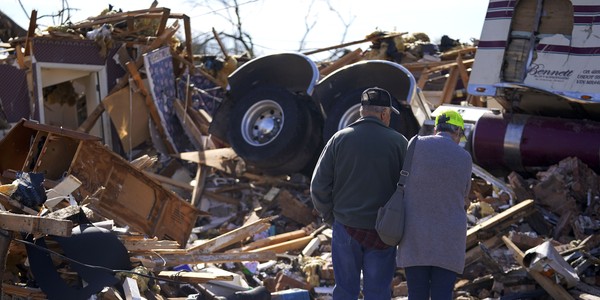 A couple stand close together, surveying extensive damage from a tornado, including a the tractor unit of a semi truck atop a pile of debris.
