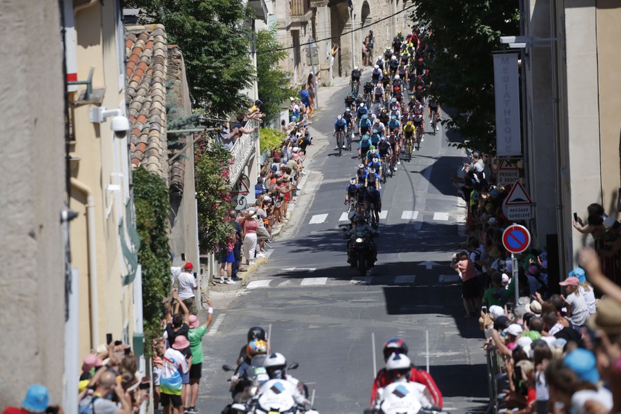 Cyclists ride down a narrow street lined with spectators.