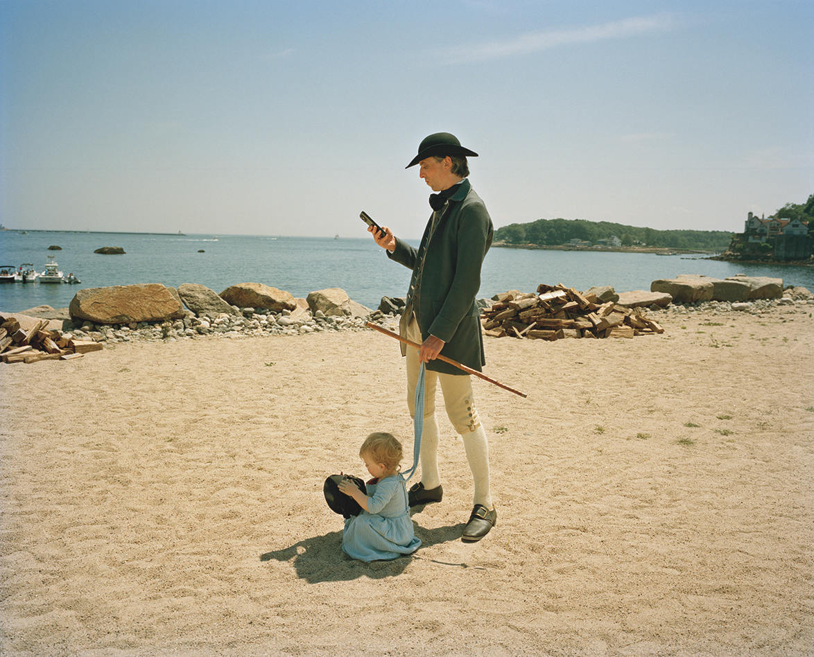 photo of male reenactor in period dress with hat, coat, breeches, and cane standing on sandy beach looking at his cell phone, with very young child playing at his feet