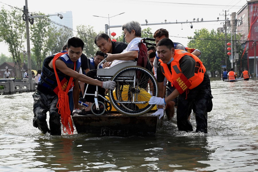 Several people work to carry a woman in a wheelchair over a flooded road.