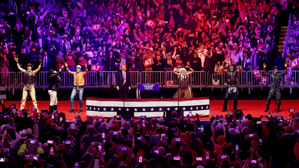Five men in different costumes dance on a big stage with another man in a suit and red tie.