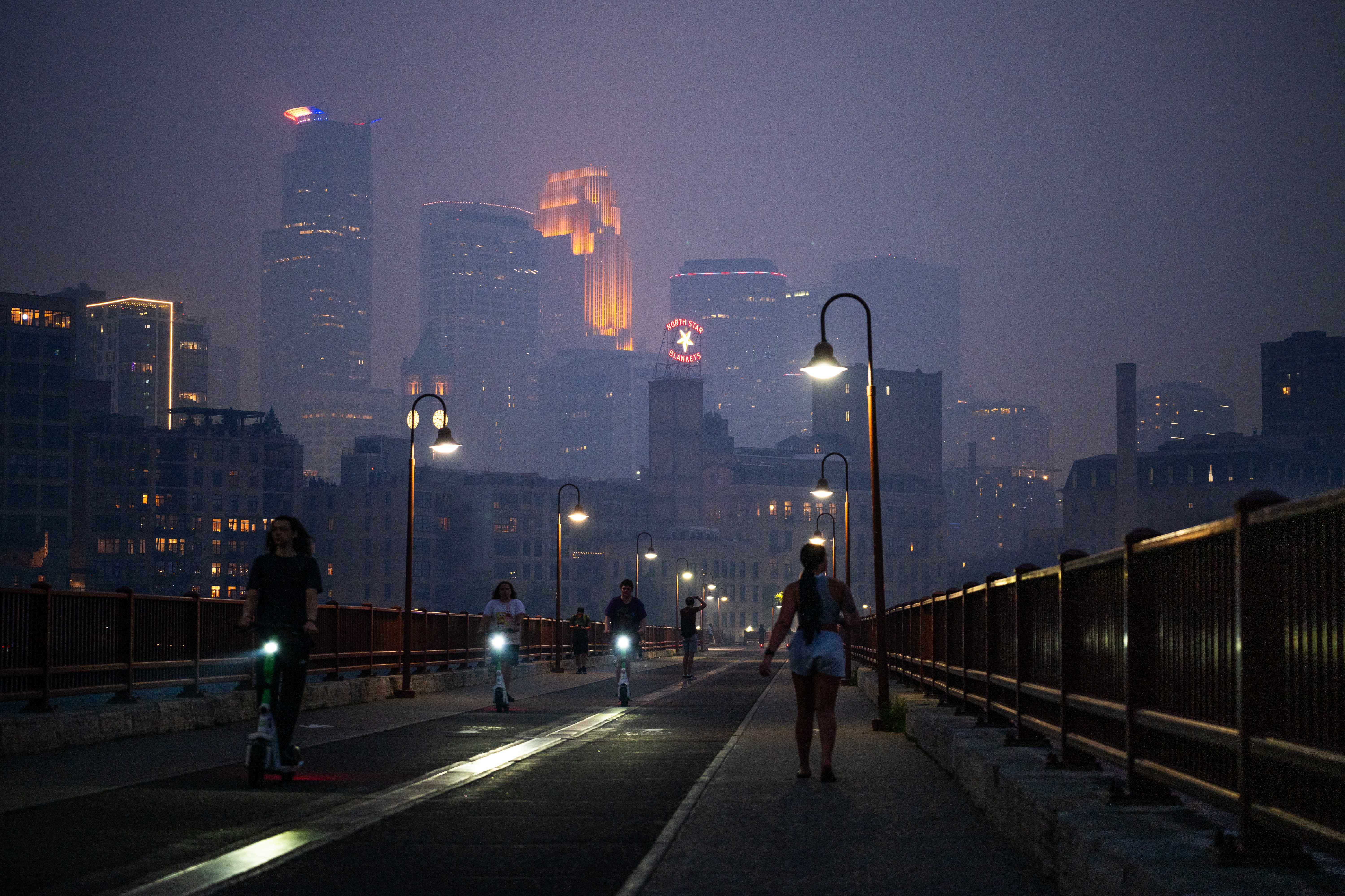People walk on a bridge at night, with tall city buildings in the background obscured by smoke.