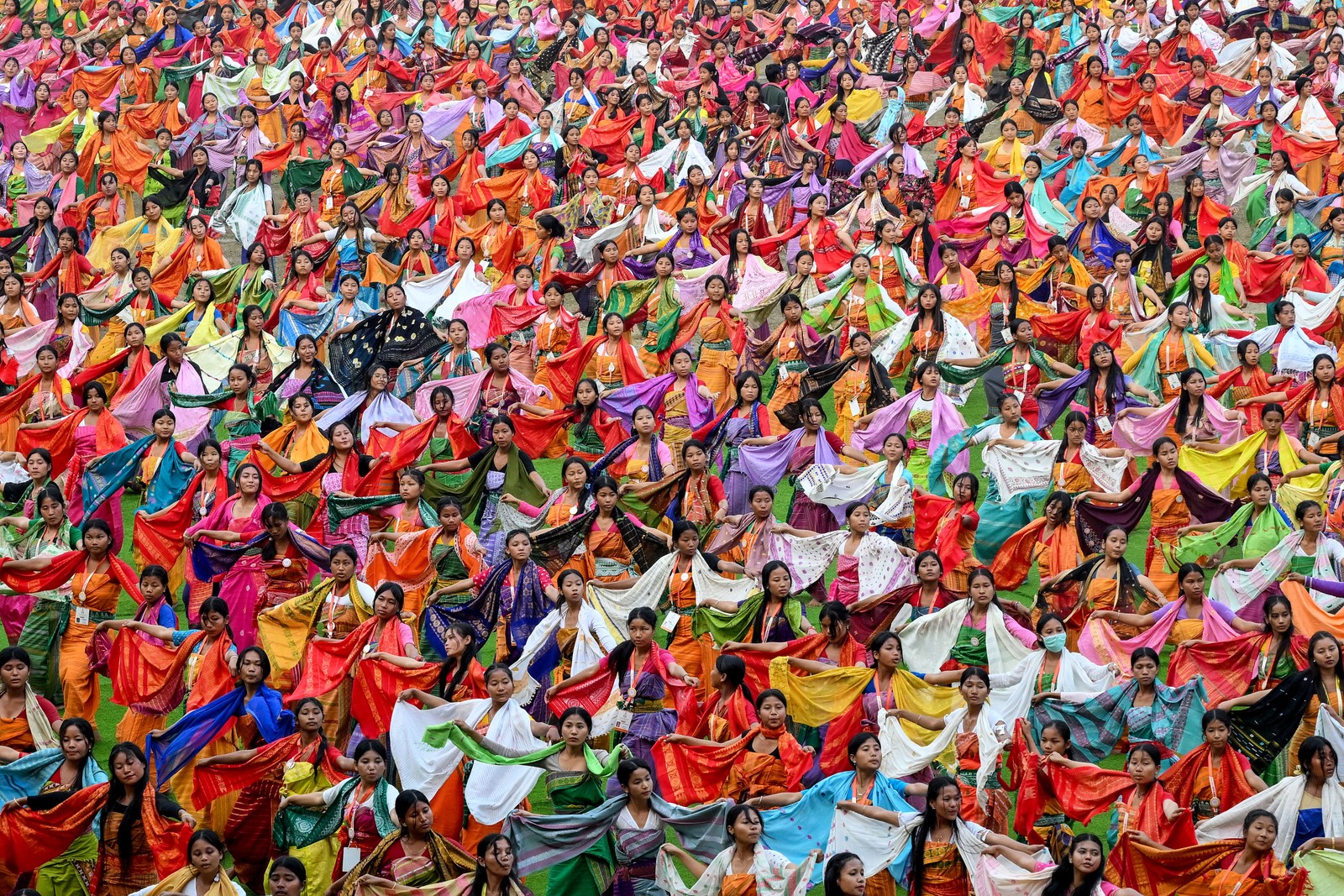 Dozens of dancers perform in unison, wearing brightly-colored scarves and dresses.