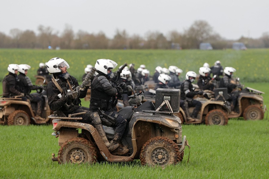 About a dozen pairs of riot-police officers ride quad bikes through a field.