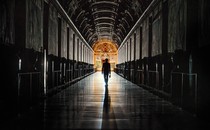Photo of a figure holding keys walking down a long, dark aisle toward a frescoed, lit door in the distance