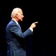 In profile, Joe Biden stands in front of a black background and points in front of him.
