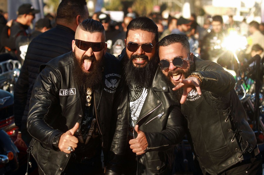 Three bearded men wearing sunglasses and leather jackets smile and make faces at the camera, with a crowd behind them.
