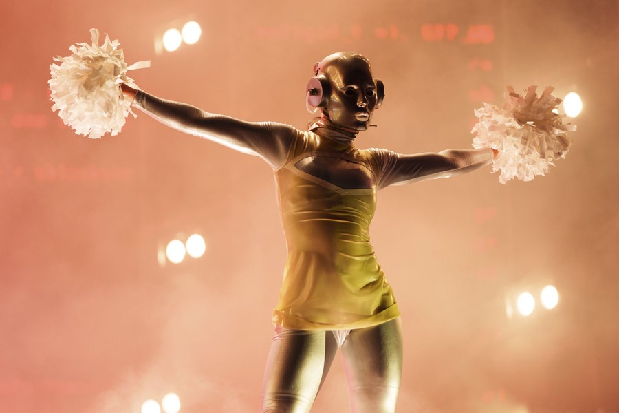 A performer wearing a robotic mask and holding pompoms appears onstage.