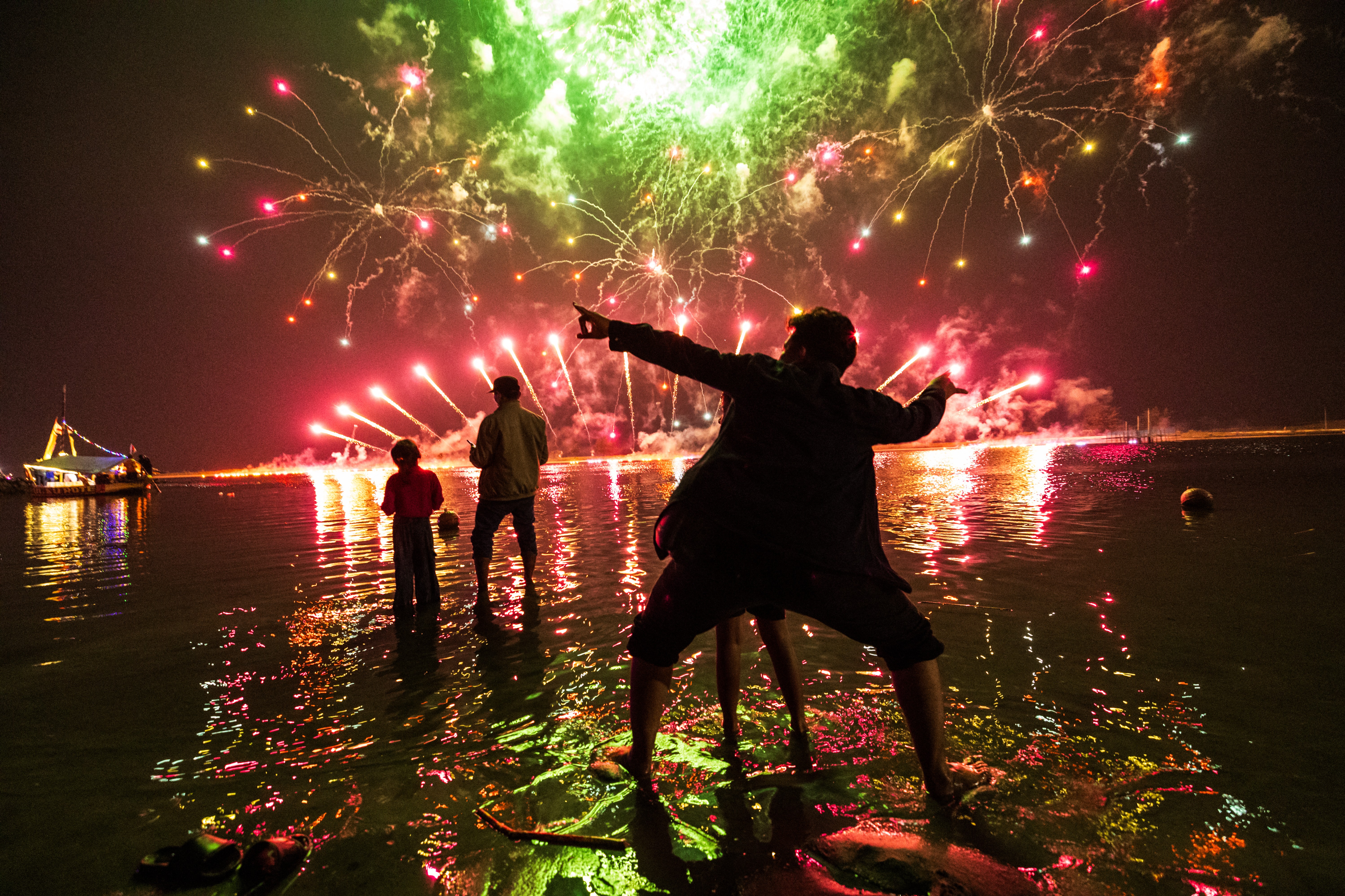 People stand in shallow water, gesturing, as they enjoy a fireworks show.