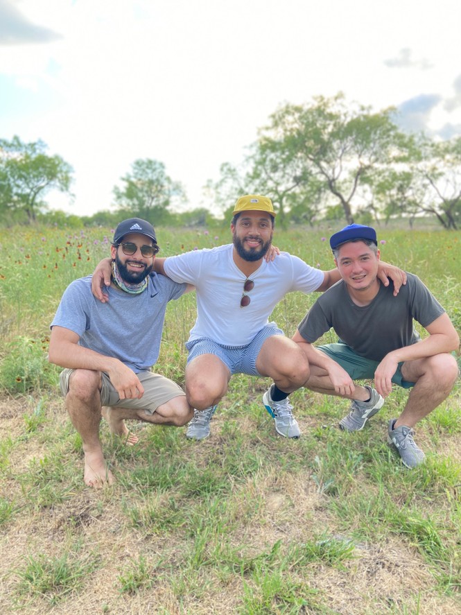 Three men crouched in a grassy field wearing shorts, t-shirts and baseball caps, with their arms around each other, smiling