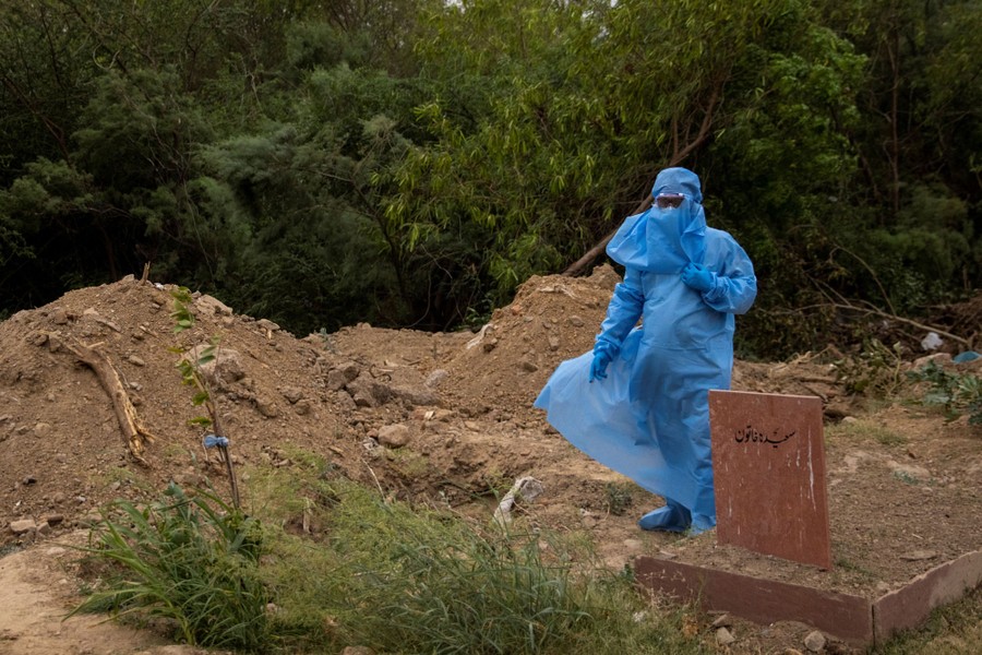 A person wearing protective gear stands near a newly-dug grave.