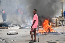 A young man stands in a city street where a fire is burning, emitting a cloud of black smoke.